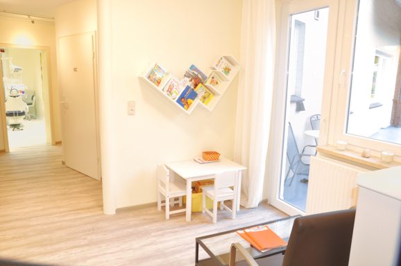 Heller kinderwartebereich mit einem kleinen Tisch und Stühlen, Regalen an der Wand und Zugang zu einem Außenbereich.